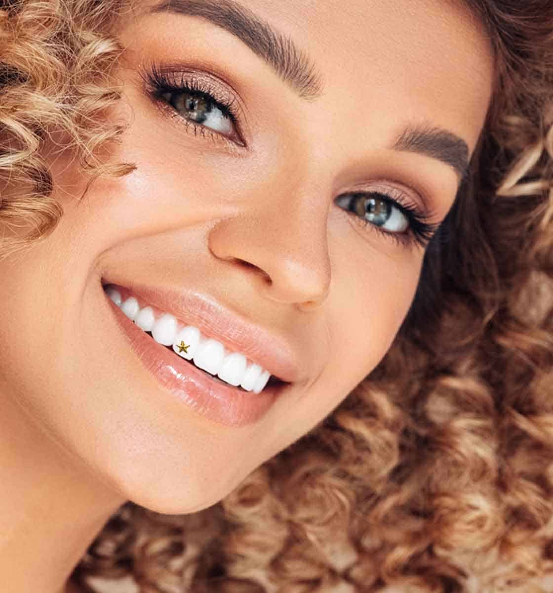 Woman with curly hair smiling, a small 24k gold star tooth gem on her upper tooth